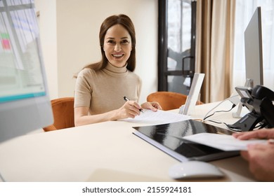 Portrait Of A Charming Caucasian Woman Holding A Pen And Filling Up A Form While Sitting At Reception Desk Of A Medical Clinic Or Financial Aid Department Office Building