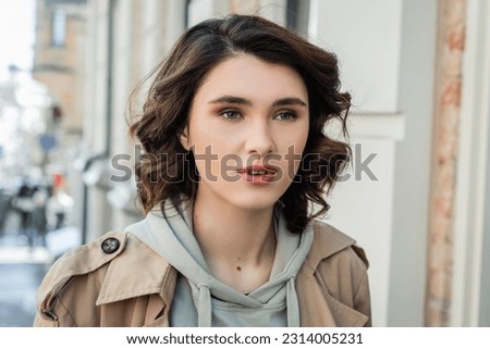 portrait of charming and fashionable woman with short brunette hair, in beige trench coat and grey hoodie looking away in European city on blurred background, street photography