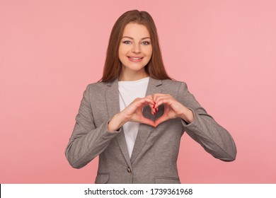 Portrait Of Charming Friendly Young Woman In Business Suit Smiling To Camera And Showing Love Heart Gesture, Symbol Of Generosity, Care And Hope. Indoor Studio Shot Isolated On Pink Background