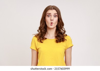 Portrait Of Charming Funny Teen Girl With Wavy Hair In Yellow T-shirt Shows Fish Face Grimace With Pout Lips, Makes Ridiculous Childish Comical Grimace. Indoor Studio Shot Isolated On Gray Background.