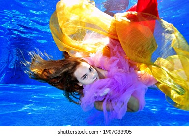 Portrait Of A Charming Girl With Long Hair Who Swims And Plays Underwater In A Pool With Yellow And Red Fabrics. Fashion Portrait. Underwater Photography. Horizontal Orientation.