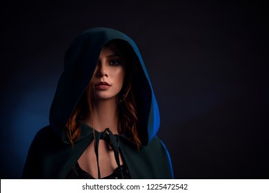 Portrait of charming and gorgeous girl with red hair looking at camera. Close up of beautiful attractive woman in emerald cloak hiding her face under hood. Concept of magic and mysterious tails.