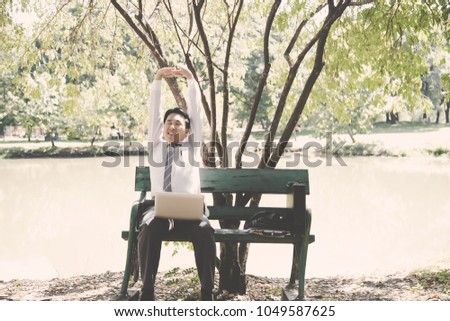Portrait charming handsome businessman. Attractive guy is sitting on bench at park in afternoon. Business man feels relaxed, happy, comfortable. He stretches his arms and back after finish working.