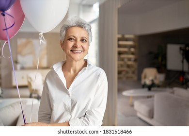 Portrait Of Charming Happy Mature Female Wearing Formal Shirt Having Fun At Her Retirement Party, Posing In Office Interior With Helium Balloons, Looking At Camera With Cheerful Broad Smile