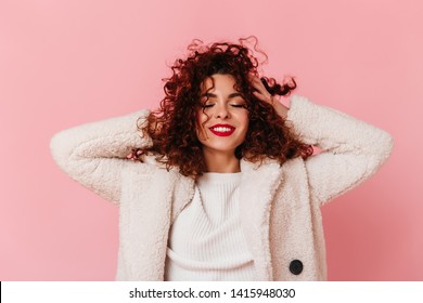Portrait of charming lady with red lipstick and snow-white smile dressed in bright eco-coat and touching her curly hair on pink background - Shutterstock ID 1415948030