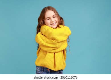 Portrait of charming little girl embracing herself and smiling from happiness, positive self-esteem, wearing yellow casual style sweater. Indoor studio shot isolated on blue background. - Shutterstock ID 2060116076