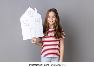 Portrait Of Charming Little Girl Wearing Striped T-shirt Holding Paper House And Smiling Friendly, Advertising Realtor Services, House Purchase. Indoor Studio Shot Isolated On Gray Background.