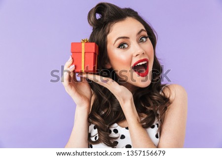 Portrait of charming pin-up woman 20s in vintage polka dot dress smiling while holding red present box isolated over violet background