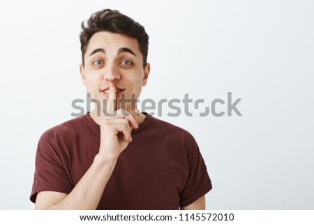 Portrait of charming playful male student in casual trendy t-shirt, lifting eyebrown with happy friendly expression, saying shh while showing shush with forefinger over mouth, smiling curiously