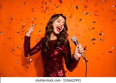 Portrait of charming pop start girl have karaoke party on her prom sing song hold microphone enjoy loud music wear maroon outfit isolated over orange color background confetti falling