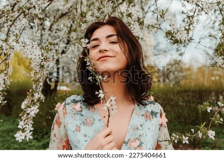 Portrait of charming pretty woman dressed flowery dress posing near apple cherry tree blossoms blooming flowers in the garden park in early spring nature. Fashion, girl model with black hair

