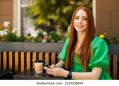 Portrait of charming redhead young woman holding mobile phone sitting at table with coffee cup in outdoor cafe terrace in sunny summer day, looking at camera, blurred background, selective focus.