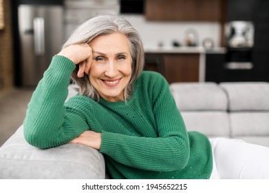 Portrait Of Charming Senior Woman With Grey Hair, Smiling Mature Female Sits On The Couch At Ease And Looks At The Camera. Beautiful Carefree Middle-aged Woman At Home