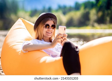 Portrait of charming smiling woman in sunglasses using cellphone and write messages to friends while lying on inflatable mattress on summer beach