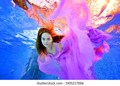 Portrait Of A Charming Teenage Girl Underwater In A Luxurious Purple Dress Against The Surreal Light From The Surface Of The Water. Underwater Concept Photography. Fashion Portrait. Vertical View.