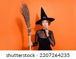 Portrait of charming witch person finger touch chin pouted lips look empty space hold broom isolated on orange color background