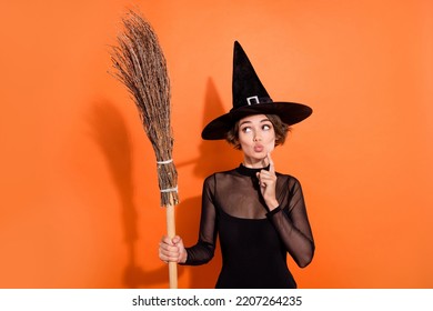 Portrait of charming witch person finger touch chin pouted lips look empty space hold broom isolated on orange color background