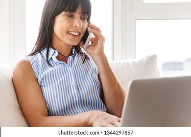 Portrait of charming woman 40s in elegant clothes using cell phone and laptop while sitting on couch in bright apartment - Powered by Shutterstock