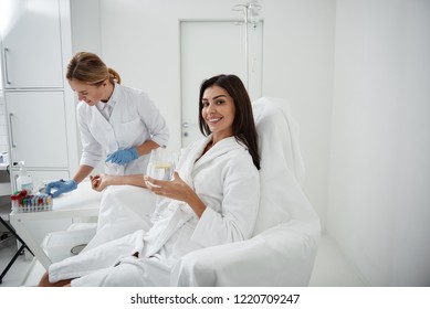 Portrait of charming woman sitting in armchair and holding glass of lemon water while doctor in sterile gloves checking vials