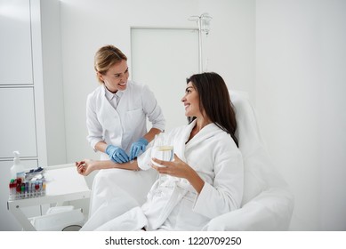 Portrait of charming woman sitting in armchair and holding glass of lemon water while doctor in sterile gloves checking IV infusion