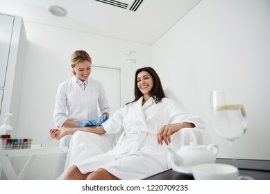 Portrait of charming woman in white bathrobe sitting in armchair and receiving IV infusion. She is looking at camera and laughing