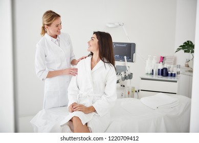 Portrait of charming woman in white bathrobe sitting on daybed and looking at beautician with smile while she touching her shoulder