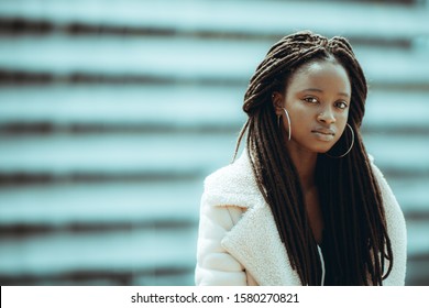 The portrait of a charming young African female in a demi-season fur coat, with long braids and big round earrings, stripes of a building facade behind her, a copy space area on the left for a message - Powered by Shutterstock