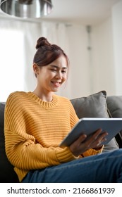 Portrait Of A Charming Young Asian Woman Relaxes In The Living Room, Sitting On Comfy Sofa And Using Portable Tablet To Watch Her Favourite Movie.