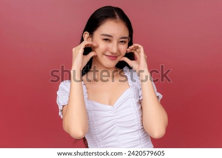 Portrait of a charming young Asian woman smiling and posing while pinching her cheeks, dressed in white with a vibrant red background.