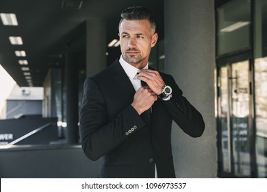 Portrait Of A Charming Young Businessman Dressed In Suit Standing Outside A Glass Building And Touching His Tie