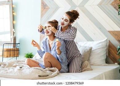 Portrait of charming young girls sitting on big bed and enjoying time together. Cute models in pyjamas posing with face masks. Youth skincare and interior concept - Powered by Shutterstock