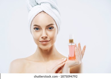 Portrait Of Charming Young Healthy Woman Holds Bottle Of Expensive Parfum, Enjoys Pleasant Aroma, Has Healthy Skin, Wears Bath Towel On Head, Stands Topless Indoor. Women And Cosmetics Concept