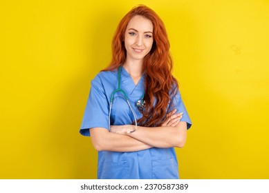 Portrait of charming young red-haired doctor woman over yellow studio background standing confidently smiling toothily with hands folded - Powered by Shutterstock