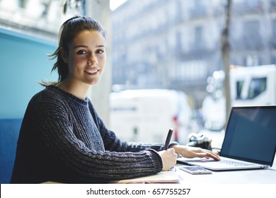 Portrait of charming young student sitting in coworking space looking at camera while making researches browsing information on laptop computer with mock up screen connected to wireless internet