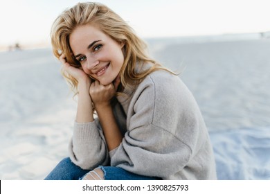 Portrait of charming young woman with blond hair, dressed in cashmere sweater. Girl sits on beach and enjoys spring day