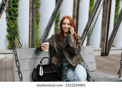 Portrait of charming young woman with long red hair talking on smartphone swinging on city swings, holding coffee cup. Happy female in brown leather jacket enjoy cellphone talking on swing alone. - Powered by Shutterstock