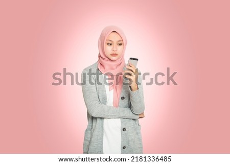 Portrait of cheeky young Asian Muslim woman looking at phone screen isolated on pink background