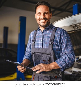 Portrait of cheerful adult man car mechanic in overalls stand in a bustling garage, hold a clipboard with a car hood open in the background, symbolizing professionalism and expertise - Powered by Shutterstock