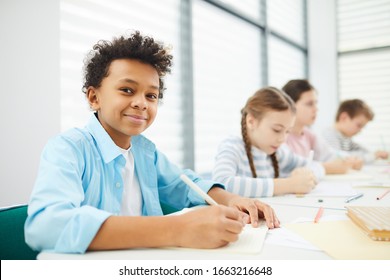 Portrait of cheerful African American boy sitting at school desk with his classmates looking at camera smiling, copy space - Powered by Shutterstock