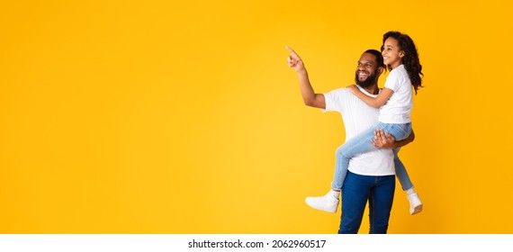 Portrait Of Cheerful African American Dad Holding Daughter On Hands, Pointing Finger Aside At Free Copy Space On Yellow Orange Background. Positive Father Showing His Child Empty Place, Panorama