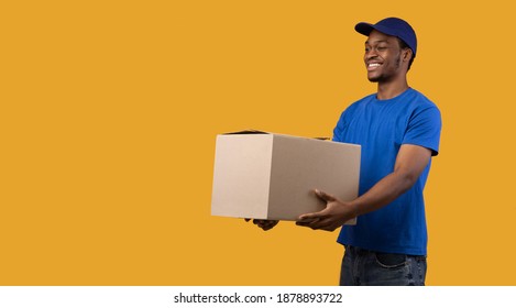 Portrait Of Cheerful African American Delivery Guy Loader Wearing Blue Cap And Uniform Holding Cardboard Container, Giving It To The Side, Copy Space, Banner. Man Isolated On Orange Studio Background