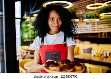 Portrait Of Cheerful African American Female Pastry Chef Smiling At Camera While Holding Big Plate With Sweet Chocolate Pastries Showing Own Desserts.Positive Confectioner With Delicious Cakes