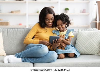 Portrait Of Cheerful African American Mom And Little Daughter Relaxing With Digital Tablet While Sitting Together On Couch In Living Room, Smiling Black Mother And Kid Using Modern Gadget At Home - Powered by Shutterstock