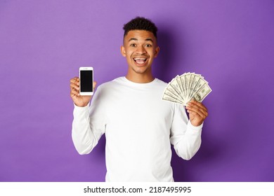 Portrait Of Cheerful African-american Man, Showing Mobile Phone Screen And Money, Standing Against Purple Background