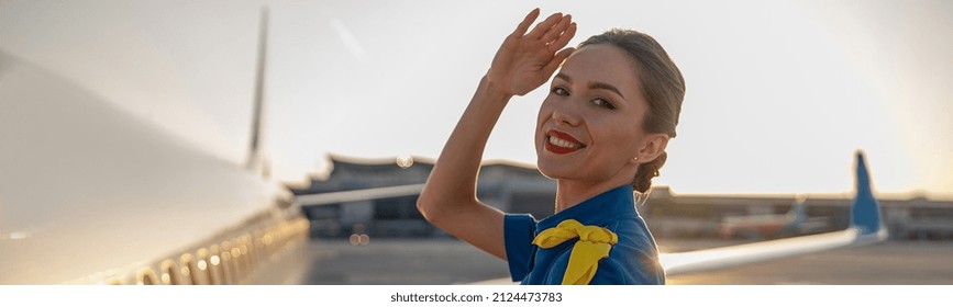 Portrait of cheerful air stewardess in blue uniform smiling at camera, posing outdoors at the sunset. Commercial airplane near terminal in an airport in the background. Aircrew, occupation concept - Shutterstock ID 2124473783