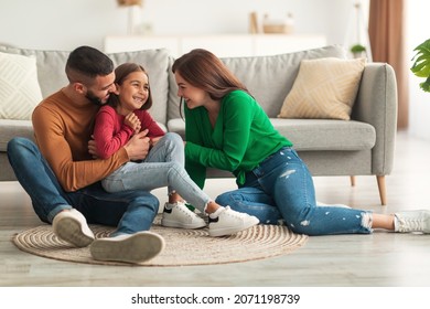 Portrait of cheerful Arab man, woman and girl playing and having fun at home together, mom amd daddy tickling laughing daughter sitting on the floor carpet in living room. Family Leisure