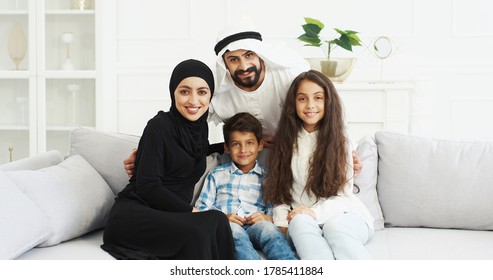 Portrait of cheerful Arabian family with two kids sitting on couch and smiling joyfully to camera. Small cute boy and girl with mother and father at home. Muslim parents with little children hugging.