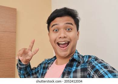 Portrait of cheerful Asian man in casual shirt taking picture of himself or selfie, making peace hand gesture in the bedroom background - Powered by Shutterstock