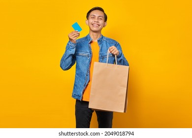 Portrait Of Cheerful Asian Man Holding Shopping Bag And Showing Credit Card Over Yellow Background