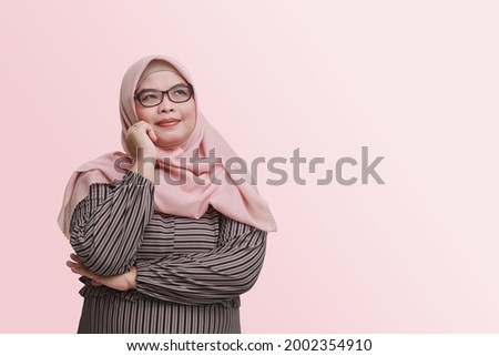 Portrait of cheerful Asian woman with hijab, standing against pink background, thinking about question with hand on chin